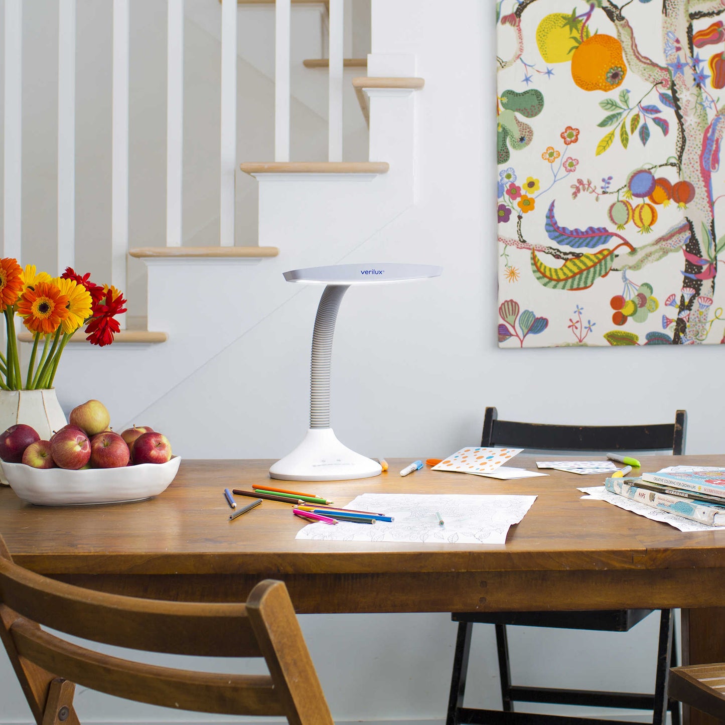 A Verilux Smartlight sitting a table lighting a child's work space. The work space has crayons and colored pencils and some coloring books. Also lots of fruit on the table and a picture on the wall.